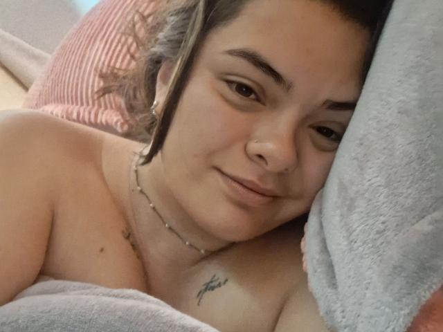 KatherinLatin with light brown skin, dark eyebrows, and a nose ring, lying down, topless, showing small tits, tattoo on right chest, smiling, grey blanket, brown hair.