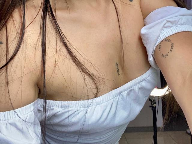 Os seios de SophiaMarx em uma blusa branca com um ombro descoberto, mostrando uma pequena tatuagem sob o seio esquerdo e outra no braço direito. Cabelo castanho longo, pele clara, foco suave..