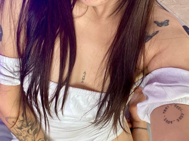 SophiaMarx met lang bruin haar, een witte top met open schouders, tatoeages op de borst en arm, vlindertatoeages op de schouder, kleine borsten, zichtbare decolleté, een tatoeage onder de borst, een tatoeage op de arm..