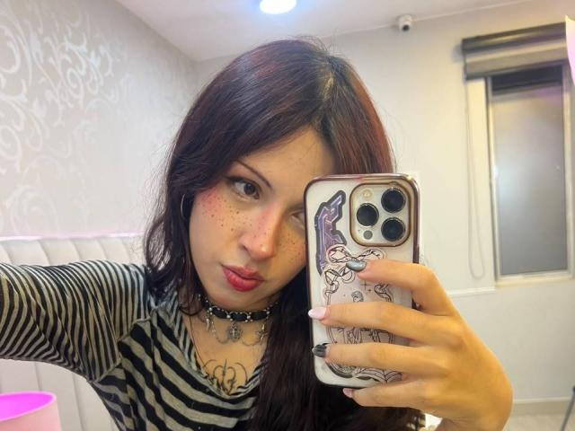 ValeryBeaumont with dark red hair, zebra-striped shirt, black choker, pink lipstick, freckles, holding phone with bunny case, taking selfie, showing tattoo on chest, pouty expression.