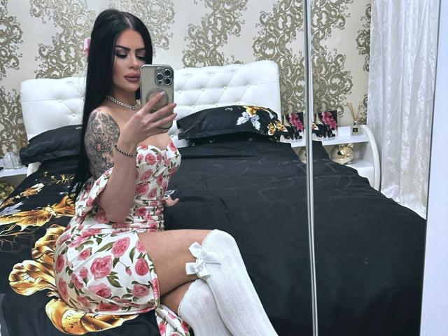 VanessaVale, con lunghi capelli neri, vestito floreale, calze bianche a ginocchio, che tiene un telefono, seduta su un letto nero, mostra un braccio tatuato e piccoli seni, mentre scatta un selfie allo specchio..