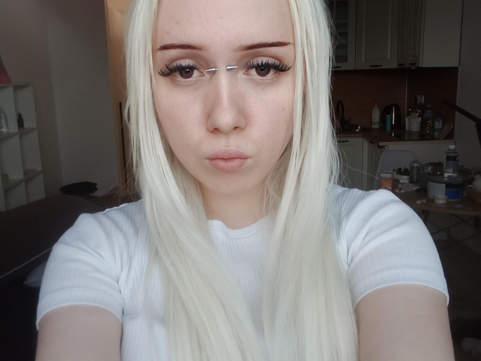 Nichole Teano, de piel clara, con cabello rubio platino largo, usando gafas, una camiseta blanca, labios fruncidos, mirando directamente a la cámara, en una cocina, con maquillaje sutil y pestañas largas..