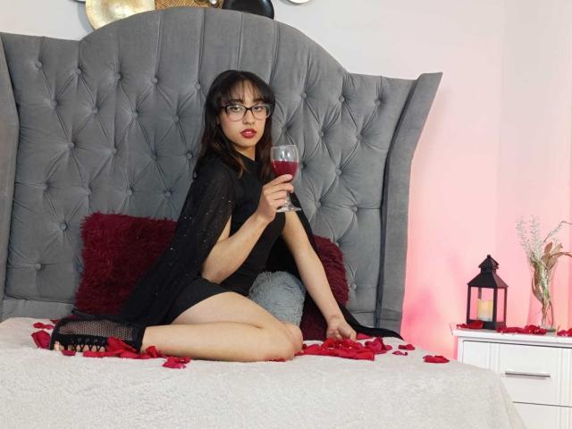 CamiVargas met een bril, een zwarte jurk en rode lippenstift, die wijn drinkt, zittend op een bed met rozenblaadjes, een grijze hoofdbord, een witte nachtkastje en een lantaarn, waarbij haar achterwerk zichtbaar is..