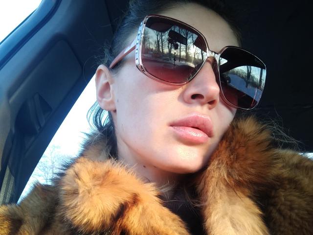 LunaBrice with shiny pink lips, wearing large brown aviator sunglasses, and a fur coat, reflecting a parking lot outside the car window. Her skin is pale and smooth.