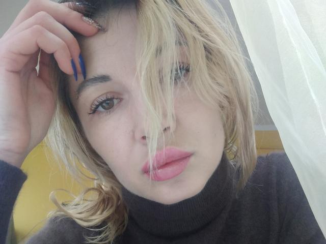 LunaBrice, com cabelo loiro, lábios rosados e unhas azuis, vestindo um cachecol marrom, tem a mão no cabelo, exibindo suas unhas longas. Pele clara e suave..
