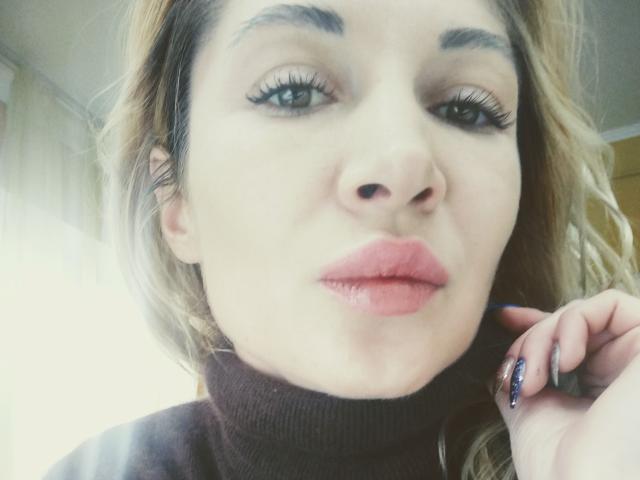 Le visage de LunaBrice, avec ses lèvres roses et pulpeuses, ses longs cils, ses cheveux blonds avec des racines foncées, son col roulé noir et son vernis à ongles violet foncé, touchant sa joue..