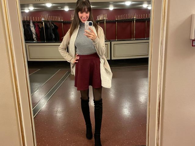Laylavon Hohensee, com um sorriso adorável, vestindo um suéter cinza de gola alta, um cardigã branco, uma saia vermelha, meias pretas e botas pretas que chegam até o joelho, tirando uma selfie no espelho..