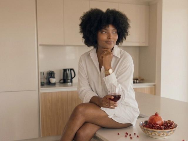 RoldanStefy, dark-skinned with afro, wearing white shirt, sitting on kitchen counter, holding red wine, wooden cabinets, pomegranate bowl with red seeds, looking off, right leg crossed.