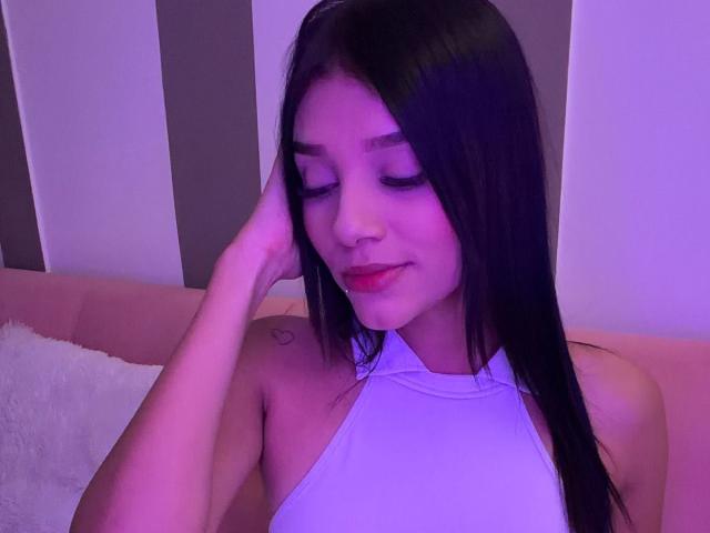 SaraMillerOne, con lunghi capelli neri, indossa un top bianco con spalline, sorride con gli occhi chiusi, ha una mano sulla testa, un piccolo tatuaggio sulla spalla destra, illuminazione rosa-viola, rossetto rosso visibile..