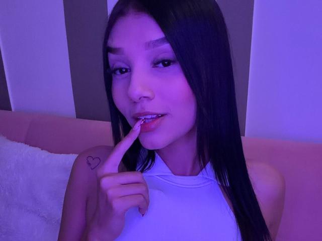SaraMillerOne, con cabello negro largo, top de tirantes blanco, dedo en los labios, iluminación morada-azul, tatuaje de corazón en el hombro izquierdo, ligera sonrisa, mirando a la cámara..