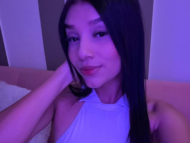 SaraMillerOne, avec de longs cheveux noirs, une peau légèrement violette et un sourire discret, porte un haut blanc à encolure dégagée et touche son cou. Ses seins sont partiellement visibles..