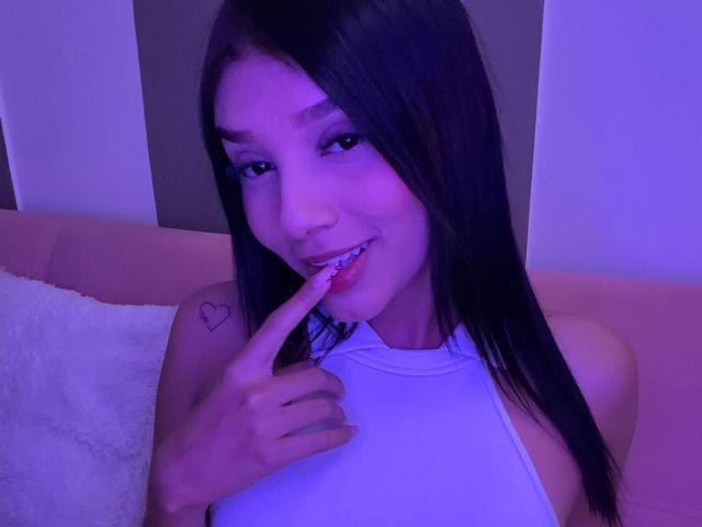 Sara Miller, una mujer de cabello negro largo, con un top blanco de tirantes y un tatuaje de corazón en el hombro derecho, mordiéndose el dedo, sonriendo bajo una luz púrpura. Sus pechos son pequeños..