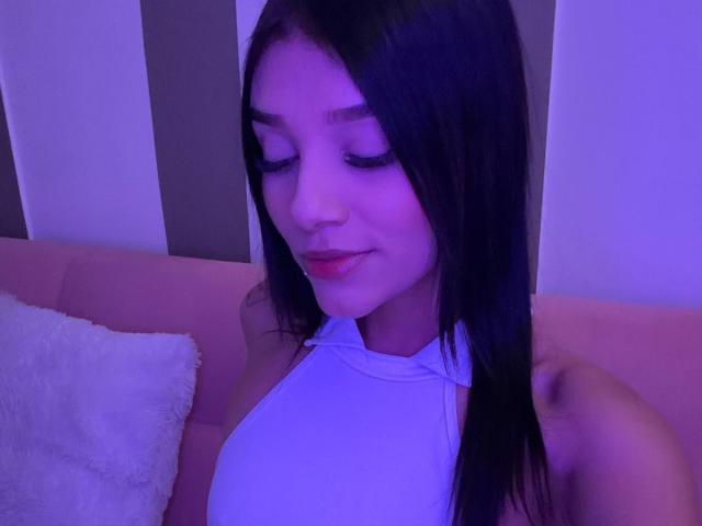 Sara Miller, com longos cabelos pretos, vestindo uma blusa branca de alças, tem os olhos fechados e um leve sorriso, sentada em um sofá rosa com uma almofada branca..