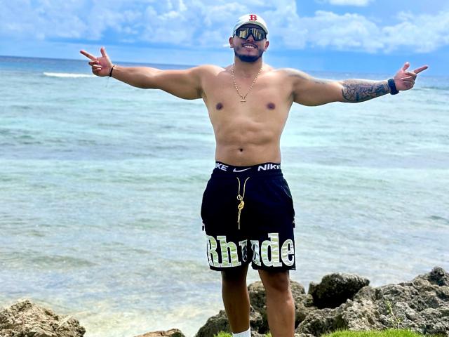Owen Pierce, sem camisa, em pé sobre rochas, com os braços estendidos, vestindo shorts pretos da Nike com a inscrição "Rhude", boné branco, óculos de sol, braço tatuado, colar e pingente, apontando com os dedos..