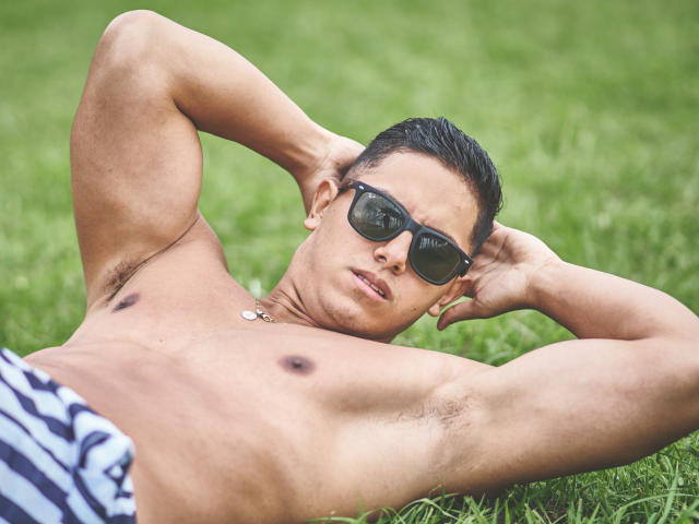 Owen Pierce, sem camisa, usando óculos de sol pretos, deitado na grama com os braços atrás da cabeça, mostrando pelos nas axilas, shorts listrados e um colar pequeno, com uma expressão séria..