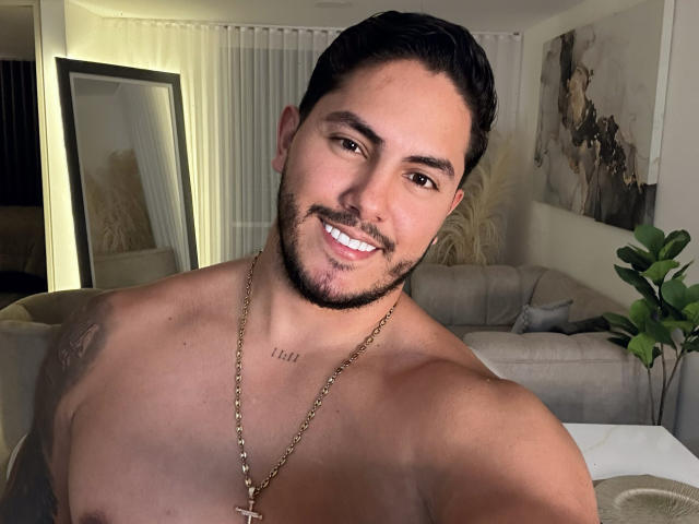 Owen Pierce, senza camicia, sorridente, capelli corti e scuri, barba, pelle abbronzata, collana d'oro, tatuaggio sul petto, muscoli tonici, occhi marroni, che guarda verso la fotocamera, soggiorno, pianta sullo sfondo..