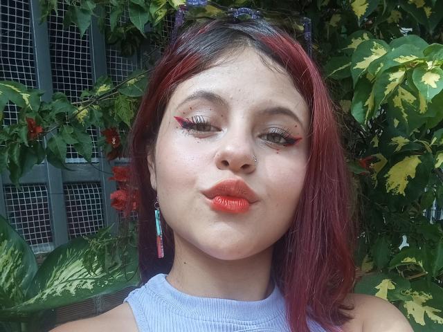 Sophie Hill a les cheveux teints en rouge, un anneau de nez et un rouge à lèvres rouge vif. Elle porte un eye-liner bleu avec des accents roses et un haut bleu clair sans manches..
