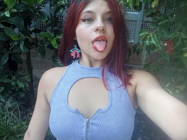 Sophie Hill, con cabello rojo, sacando la lengua, vistiendo una camiseta azul texturizada con un corte y cremallera, aretes y un piercing en la nariz, sosteniendo un selfie..