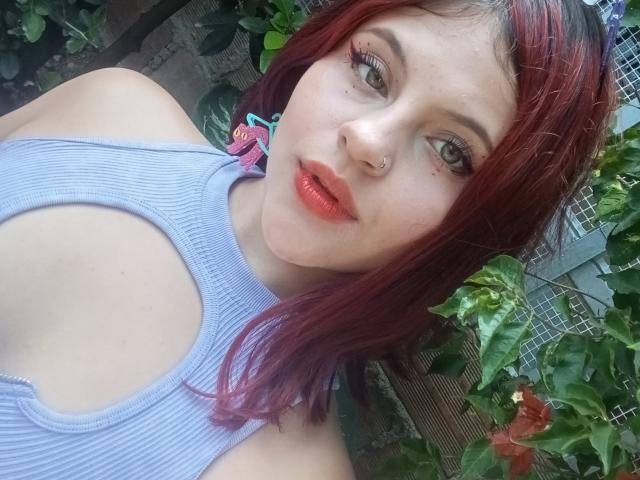 Sophie Hill, con cabello rojo, piercing en la nariz y un top azul a rayas, que deja al descubierto sus hombros y pecho, tiene labios rosados y brillantes y una expresión linda, ligeramente tímida..