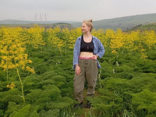 Laila Lafreniere è in un campo verde con fiori gialli, indossa una camicia blu, un reggiseno nero, una cintura rosa e pantaloni verdi, e guarda di lato con un leggero sorriso..