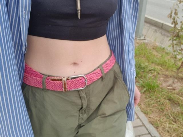 LailaLafrenieres Oberteil zeigt einen schwarzen Top, einen pinken Gürtel mit goldener Schnalle, grüne Hosen und ein blau gestreiftes Hemd. Ihr Bauchnabel ist sichtbar..
