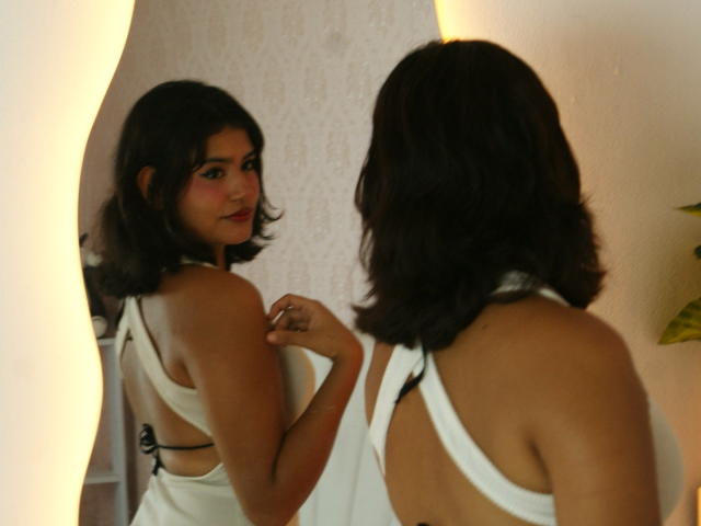 Lilith Jones, com cabelo preto curto, vestindo um vestido branco de alças, olhando para o seu reflexo em um espelho. Ela tem seios pequenos, uma figura esguia e um leve sorriso..