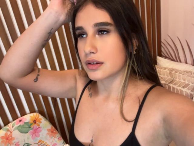 KatieDesirX met lang bruin haar, een zwart tanktopje, kleine borsten, een armtattoo, oorbellen en een tepel piercing, kijkt in de camera met haar hand in haar haar..