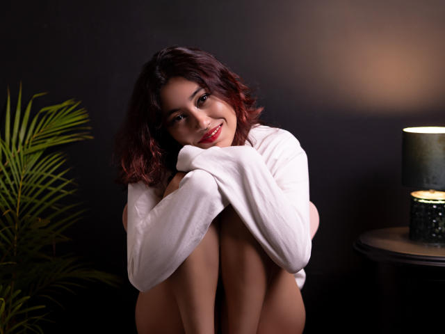 AliCipher with wavy brown hair, smiling, hugging knees, wearing a white shirt, sitting next to a green plant and black lamp, showing her bare ass.