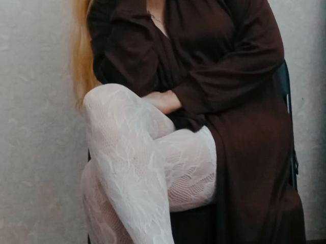 CarlyTreat è seduta con le gambe incrociate, indossa un vestito marrone e collant bianchi in pizzo, mostrando glutei e cosce, con le braccia incrociate e lunghi capelli biondi, seduta su una sedia nera..