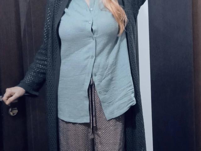Carly, avec ses cheveux blonds, portant une chemise bleue à boutons, un cardigan noir à motifs et un pantalon marron à pois, se tient près d'une porte sombre, avec sa main sur la poignée de la porte..