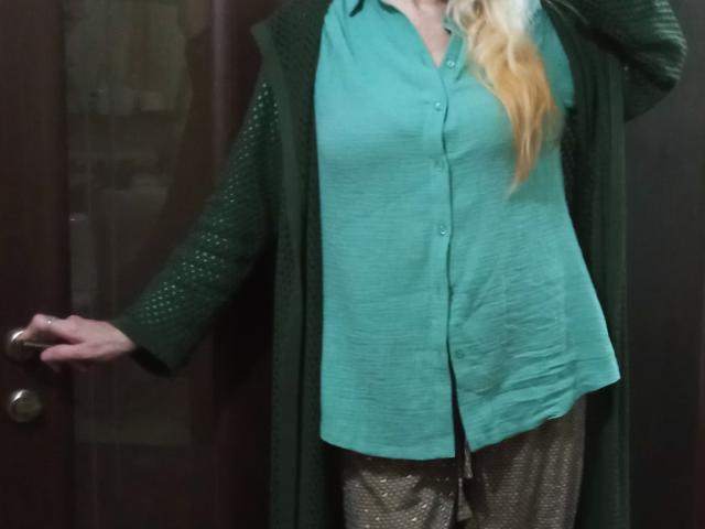 Carly, con i capelli biondi, indossa una camicia verde a bottoni, un cardigan verde e pantaloni scintillanti, ed è in piedi accanto a una porta scura, con la mano sulla maniglia..