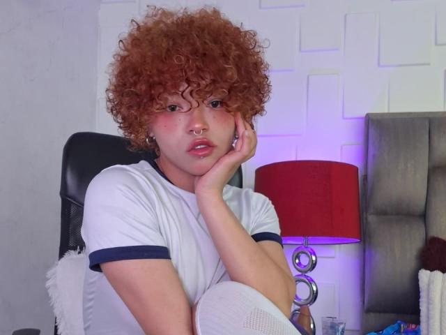 KeiryGinger with curly red hair, white tee, and nose ring, sitting on black chair, hand on cheek, looking pouty, red lamp with purple glow on right.