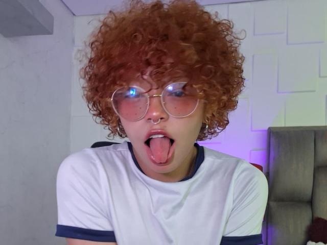 KeiryGinger with curly red hair, glasses, and nose ring, sticking out tongue, wearing a white tee with navy trim. They're looking goofy with a playful expression.