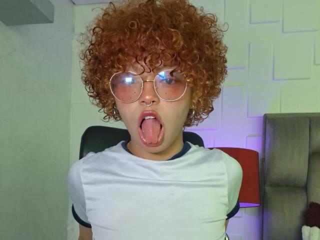 KeiryGinger with curly red hair, round glasses, and white shirt, sticking out their tongue. Their ass is not visible. KeiryGinger has freckles and a cute, surprised face.