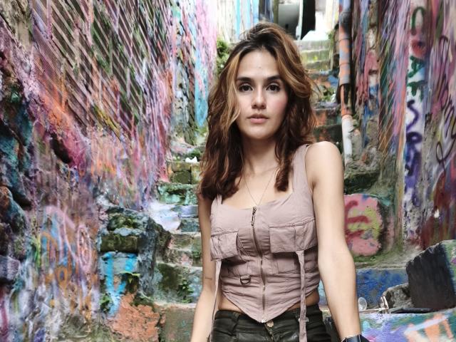 Amelie Lautrec tem cabelos castanhos ondulados, veste uma blusa cropped rosa com botões, que mostra seios pequenos, e está em uma viela colorida, coberta de grafites, usando calças verdes e com uma expressão séria..