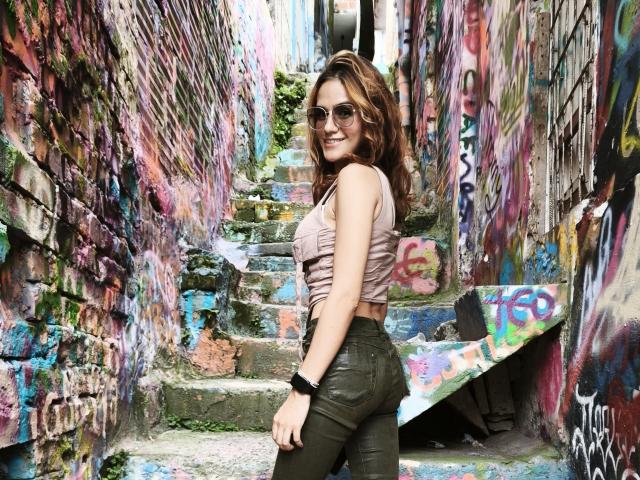 Amelie Lautrec tem cabelos castanhos cacheados, usa óculos de sol, uma blusa cropped rosa e calças de couro verde, está em pé em degraus cobertos de grafite, sorrindo, e usa uma pulseira preta..