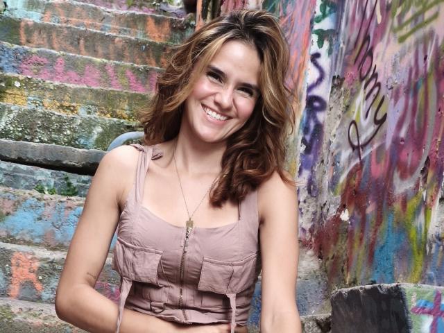 Amelie Lautrec tem cabelos castanhos ondulados, está sorrindo mostrando dentes brancos, veste uma regata rosa que revela seios pequenos, e usa um colar, estando em pé em degraus coloridos cobertos de grafite..