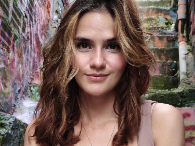 Amelie Lautrec tem cabelos castanhos ondulados com mechas loiras, um sorriso discreto e um rosto bonito. Ela está vestindo uma blusa rosa sem mangas e olhando ligeiramente para o lado..