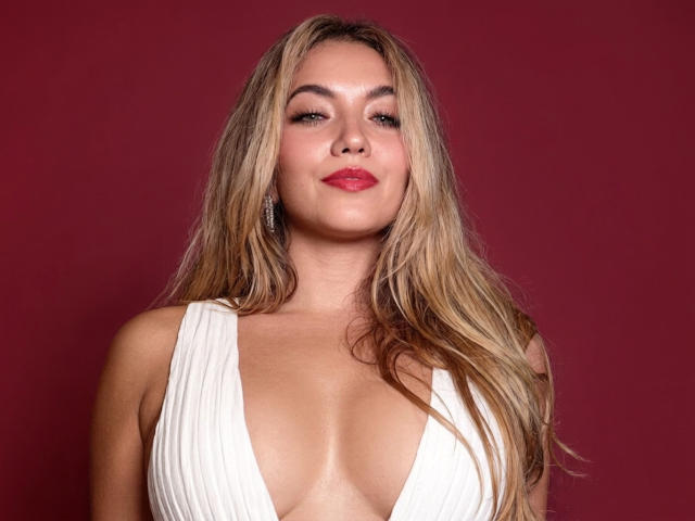 SofieWhite with long blonde hair, red lipstick, and white deep V-neck dress, showing her big boobs and confident smile against a solid red background.