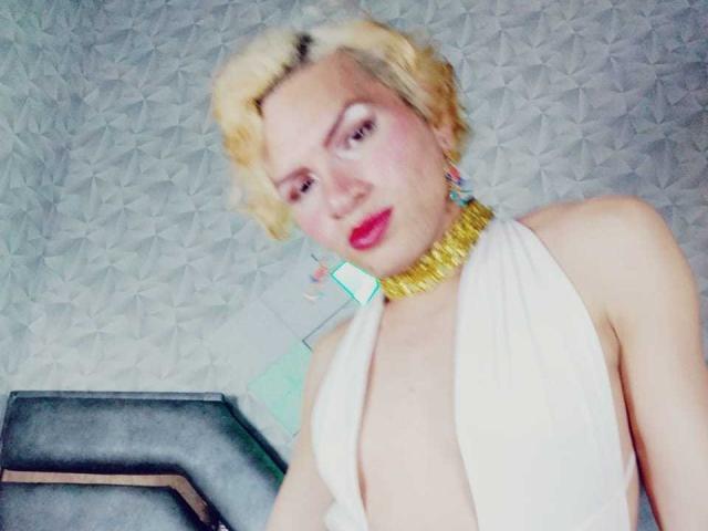 Marilyn Marios mit blonden Haaren, rotem Lippenstift und einer Goldkette, trägt ein weißes Top mit Halstuch, das ihre Brust freilegt, und blickt in die Kamera..