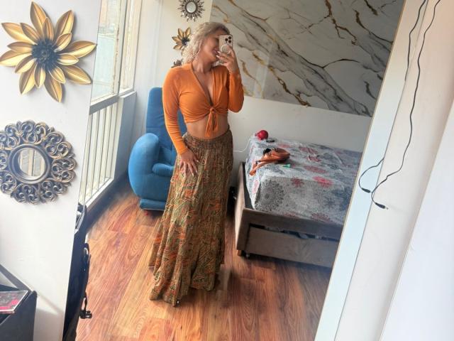Flora Saenz, avec ses cheveux blonds bouclés, son haut orange à manches longues et sa jupe à motifs, se tient dans une chambre, tenant un téléphone, prenant un selfie devant un miroir. On voit une chaise bleue, un sol en bois et un mur en marbre..