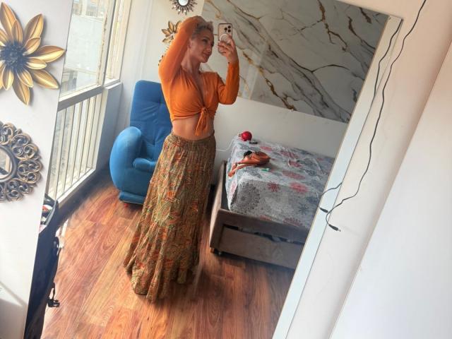 Flora Saenz, portant un crop top orange noué sur le devant, laissant entrevoir ses petites seins, et une longue jupe fluide à motifs, se prend en photo dans un miroir, debout sur un plancher en bois..