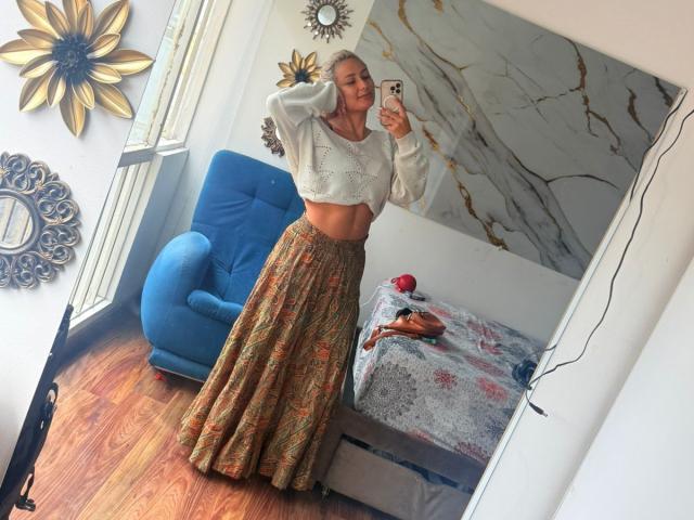 FloraSaenz pose pour une photo en utilisant un miroir, elle porte un débardeur blanc et une longue jupe à motifs, mettant en valeur son ventre plat et ses cheveux blonds, avec une chaise bleue à proximité..
