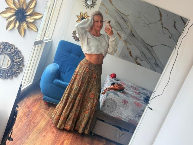 Flora Saenz, avec ses cheveux blonds, son crop top blanc et sa jupe colorée, tenant un téléphone, debout dans une pièce, avec une chaise bleue, un décor mural en marbre, un sol en bois, un selfie pris dans un miroir, souriante..