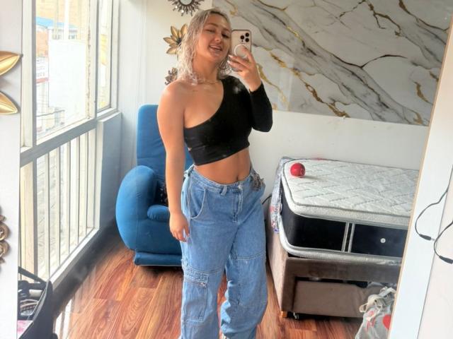 Flora Saenz, met blond haar, een zwart top met één schouder, en blauwe jeans, lacht in een spiegelzelfportret, waarbij haar getoonde buik en billen te zien zijn, terwijl ze op een houten vloer staat..