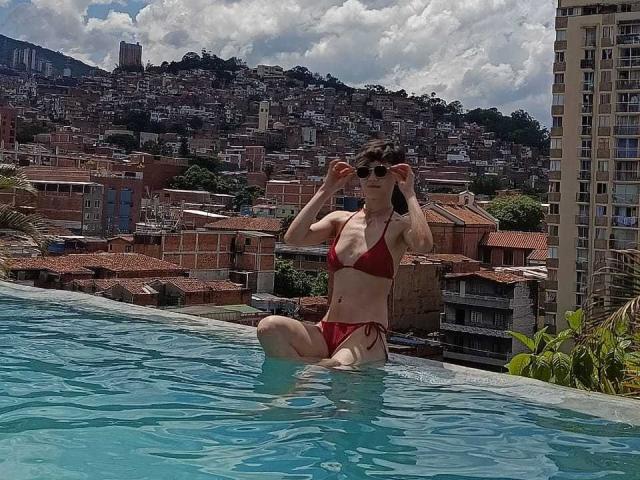 LissyLuxe, in a red bikini, sits in a rooftop pool, adjusting her sunglasses, with her tits and ass partially visible, against a city skyline.