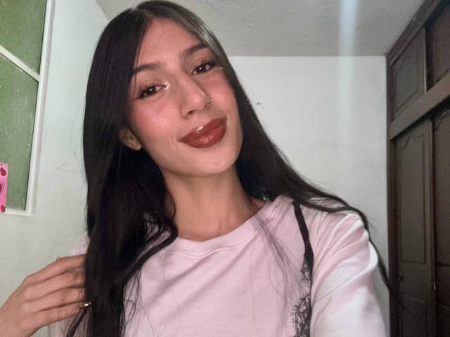 Sophie Beaumont, con cabello largo y oscuro, vestida con una camisa rosa, sonriendo con un lápiz labial rojo brillante, sosteniendo su cabello a la izquierda, en una habitación con poca luz..