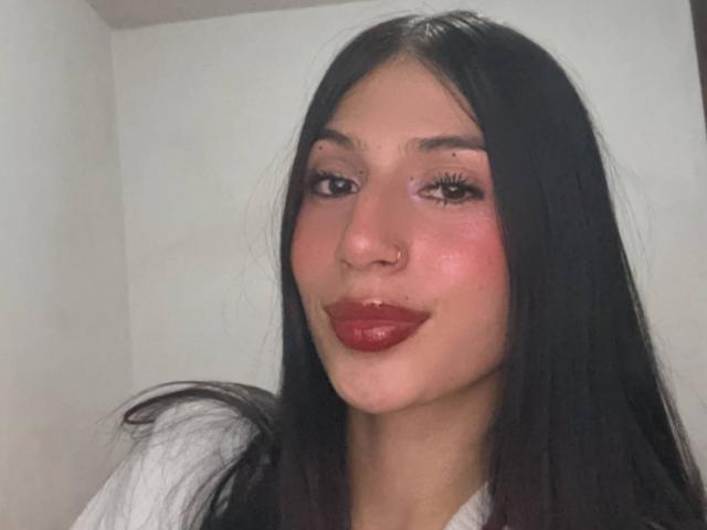 Sophie Beaumont tem cabelos pretos longos, um piercing no nariz e lábios vermelhos brilhantes. Sua pele é levemente brilhante e ela está vestindo uma camisa branca. Seus olhos estão semicerrados..