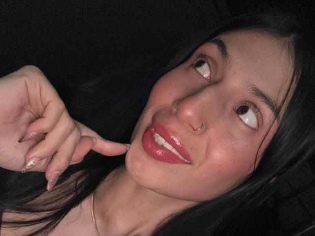 O rosto de Sophie Beaumont aparece em uma selfie, com cabelo preto, batom vermelho, um piercing no nariz e um sorriso divertido, enquanto ela toca o lábio com o dedo. Fundo escuro..