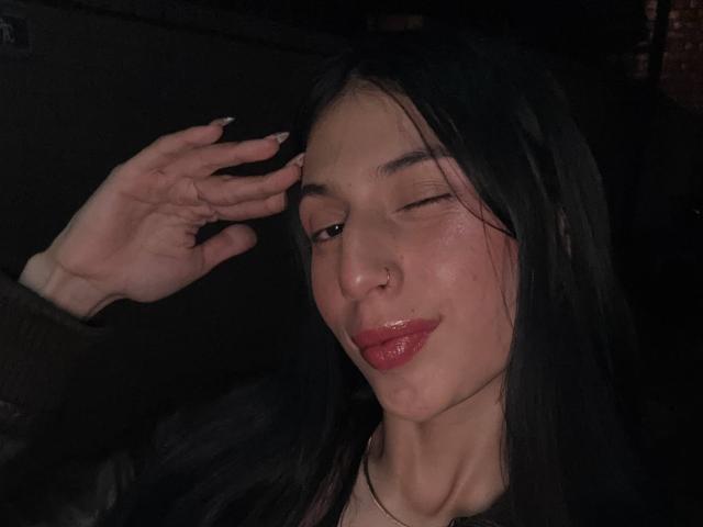 Sophie Beaumont, com cabelo preto, piercing no nariz e lábios vermelhos brilhantes, piscando, segurando a testa, mostrando um leve sorriso, vestindo uma blusa escura, em luz fraca..